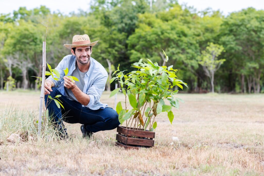 A farmer in a straw hat smiles while holding a young plant in a field.